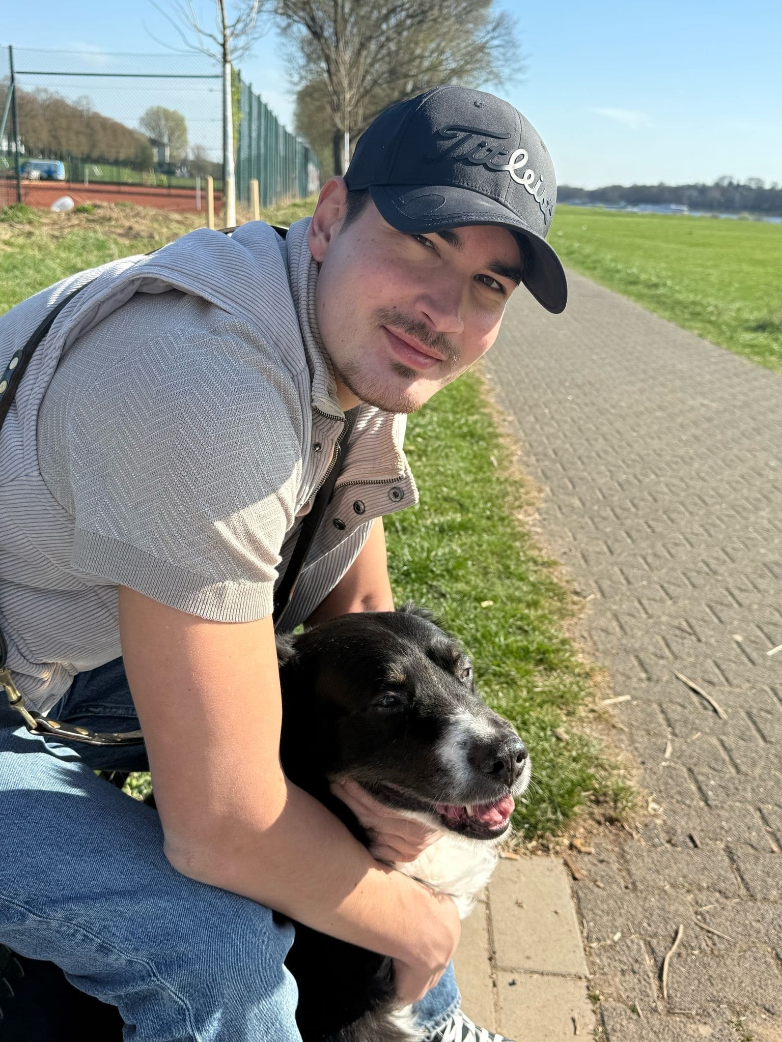 Marvin Alberg und sein Hund Yoshi sitzen auf einer Mauer bei einem Sportplatz sowie einer Wiese.